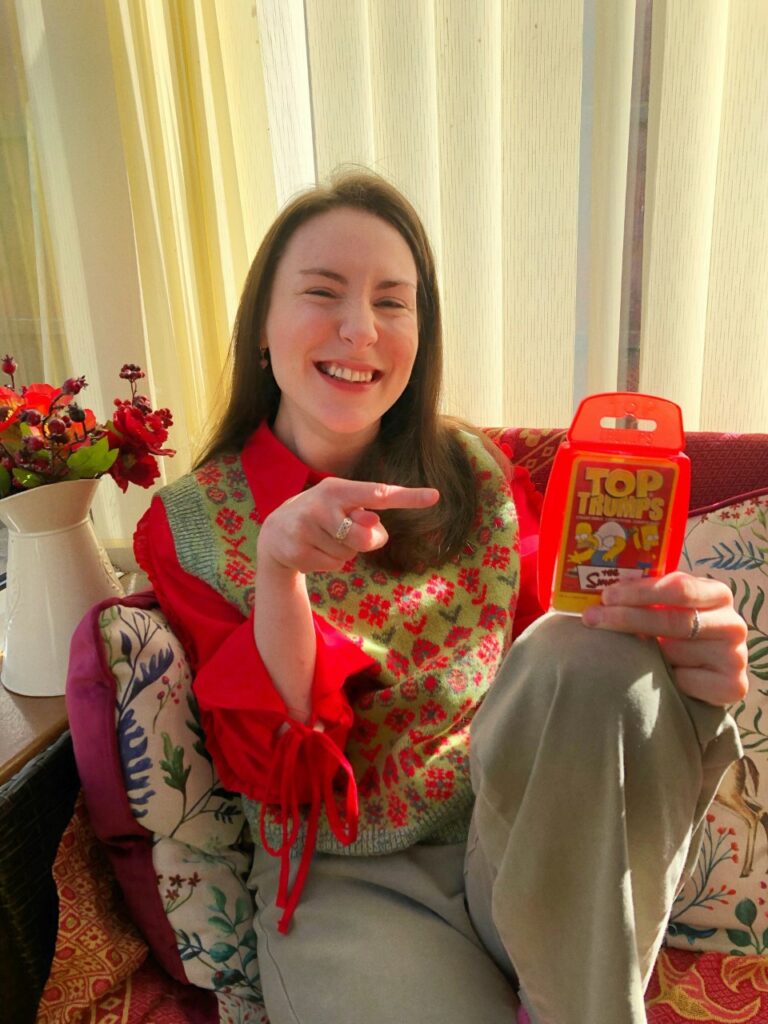Rebecca a white woman with long brown hair. is sitting on a sofa holding a box of The Simpsons Top Trump cards