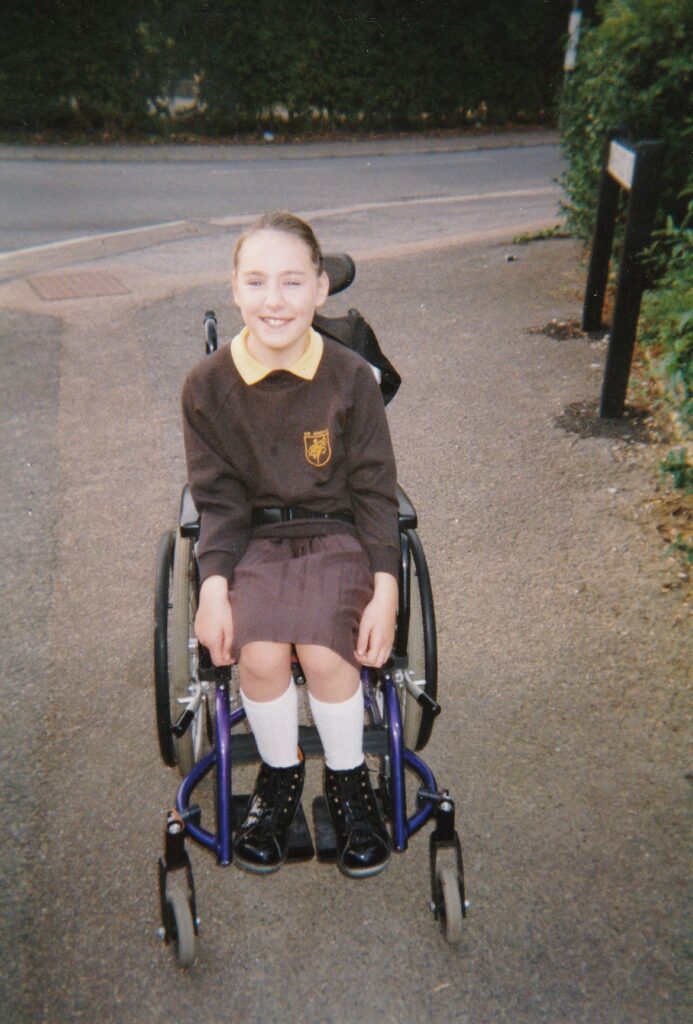An old photo of ten year old Rebecca, a white girl with brown hair, She is wearing her school uniform with Piedro boots and is sitting in her wheelchair. 